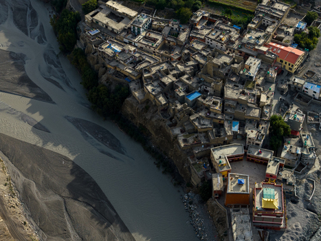 Aerial view of a traditional village with stone buildings and flat rooftops situated along a wide rocky riverbed in the mountainous landscape of Nepal.
