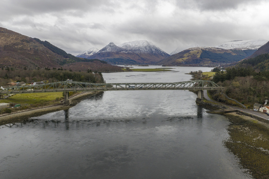 Aerial view of Ballachulish Bridge spanning Loch Leven with snow capped mountains and a cloudy sky in the background Ballachulish Scotland United Kingdom.