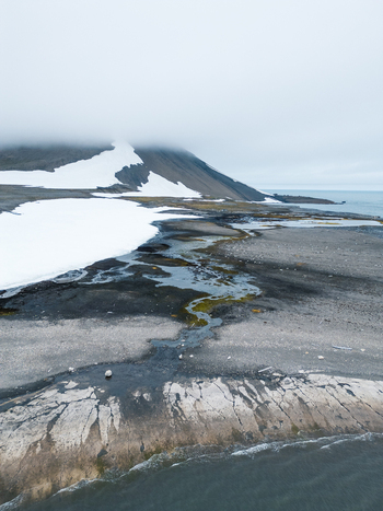 Svalbard and Jan Mayen 28 July 2025 Aerial view of the rugged arctic coastline with snow patches dark rocky terrain and a mountain peak shrouded in low clouds. Svalbard and Jan Mayen 28 July 2025 Aerial view of the rugged arctic coastline with snow patches dark rocky terrain and a mountain peak shrouded in low clouds.
