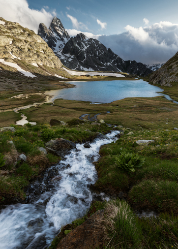 View of a rushing mountain stream flowing into a serene alpine lake surrounded by snow capped rocky peaks under a cloudy sky in the Aosta Valley Italy. View of a rushing mountain stream flowing into a serene alpine lake surrounded by snow capped rocky peaks under a cloudy sky in the Aosta Valley Italy.