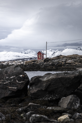 View of a small red wooden hut on a rocky shoreline with snow capped mountains under an overcast sky in Tromso Troms Norway.