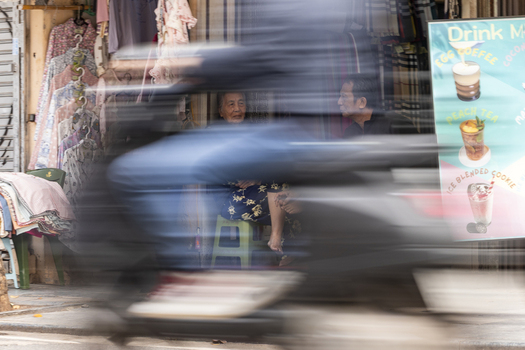 Hanoi Vietnam 11 November 2025 View of two people sitting in a shop doorway behind a blurred motorbike passing by on a busy street in Hanoi.