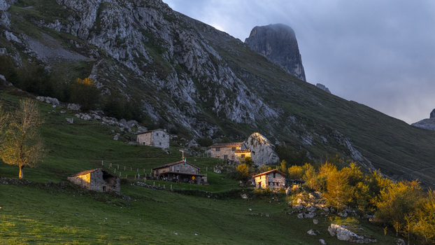 Aerial view of rustic stone houses nestled on a vibrant green hillside beneath jagged peaks illuminated by the warm glow of sunset Picos de Europa Asturias Spain. Aerial view of rustic stone houses nestled on a vibrant green hillside beneath jagged peaks illuminated by the warm glow of sunset Picos de Europa Asturias Spain.