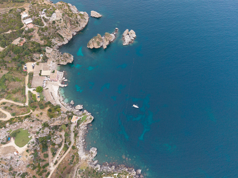 Aerial view of the rugged coastline of Sicily with turquoise waters rocky cliffs a small beach with buildings and a lone boat floating in the sea in Palermo Sicily Italy. Aerial view of the rugged coastline of Sicily with turquoise waters rocky cliffs a small beach with buildings and a lone boat floating in the sea in Palermo Sicily Italy.