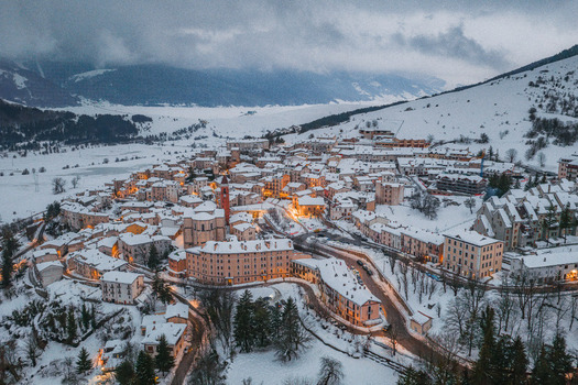 Aerial view of the historic town of Rivisondoli covered in a thick blanket of snow with warm glowing lights from buildings under a cloudy winter sky in Rivisondoli Abruzzo Italy.