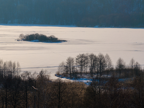 Aerial view of a frozen lake covered in snow with small islands and bare trees under soft winter light in Lithuania. Aerial view of a frozen lake covered in snow with small islands and bare trees under soft winter light in Lithuania.