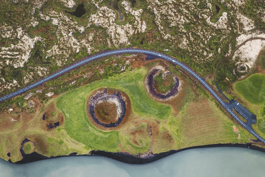 Aerial view of pseudo craters and winding road contrasting against the lush green landscape and the blue waters of Lake Myvatn Iceland.