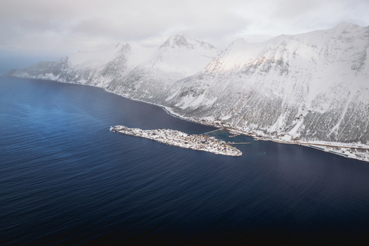 Aerial view of a tranquil island village nestled between the deep blue fjord and snow covered mountains a serene landscape of contrasting colors and textures Fjordgård Troms Norway. Aerial view of a tranquil island village nestled between the deep blue fjord and snow covered mountains a serene landscape of contrasting colors and textures Fjordgård Troms Norway.
