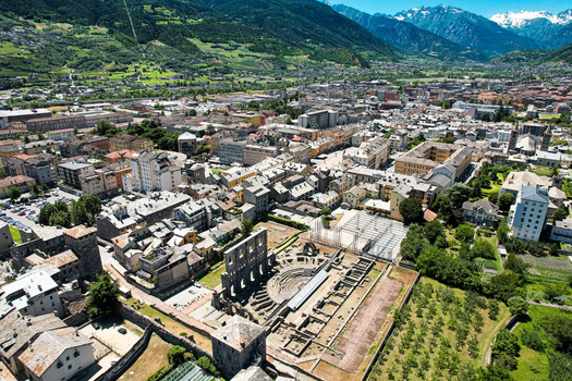 Aerial view of the ancient Roman Theatre ruins surrounded by the historic city rooftops and the majestic snow capped Alps in Aosta Aosta Valley Italy. Aerial view of the ancient Roman Theatre ruins surrounded by the historic city rooftops and the majestic snow capped Alps in Aosta Aosta Valley Italy.