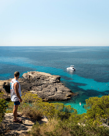Balearic Islands Spain 15 September 2023 View of a man standing on a cliff overlooking the turquoise waters of a bay in Mallorca with a small boat and swimmers. Balearic Islands Spain 15 September 2023 View of a man standing on a cliff overlooking the turquoise waters of a bay in Mallorca with a small boat and swimmers.