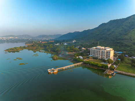 Aerial view of a hotel on the edge of a lake with a long pier extending into the water surrounded by lush trees and rolling hills under a clear blue sky in Udaipur Rajasthan India. Aerial view of a hotel on the edge of a lake with a long pier extending into the water surrounded by lush trees and rolling hills under a clear blue sky in Udaipur Rajasthan India.