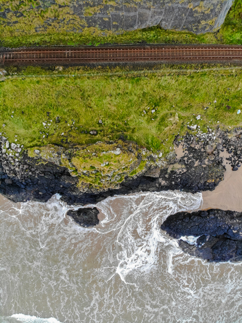 Aerial view of Mussenden Temple perched on the cliff edge above the Atlantic Ocean with a railway line running along the grassy coastline in Coleraine Northern Ireland United Kingdom. Aerial view of Mussenden Temple perched on the cliff edge above the Atlantic Ocean with a railway line running along the grassy coastline in Coleraine Northern Ireland United Kingdom.