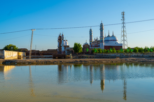 View of a mosque with ornate minarets and white domes reflected in a calm pool of water surrounded by green flags and a communication tower in Jacobabad Sindh Pakistan.