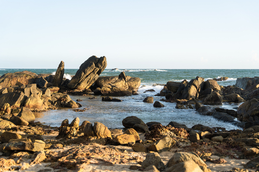 View of large dark rock formations and boulders scattered along the sandy shoreline with ocean waves crashing against the coast under a clear sky in Galle Southern Province Sri Lanka.