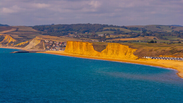 Aerial view of the golden sandstone cliffs at West Bay featuring the rugged coastline blue sea and beach huts in West Bay England United Kingdom. Aerial view of the golden sandstone cliffs at West Bay featuring the rugged coastline blue sea and beach huts in West Bay England United Kingdom.