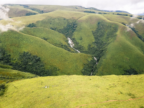 Aerial view of vibrant green rolling hills cascading into a forested valley with a glistening waterfall Obudu Cross River Nigeria.