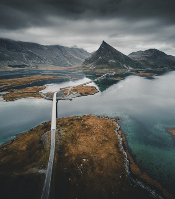Aerial view of Flakstadbruene bridge connecting islands with rugged terrain dark water and dramatic mountains under a cloudy sky in Flakstad Nordland Norway. Aerial view of Flakstadbruene bridge connecting islands with rugged terrain dark water and dramatic mountains under a cloudy sky in Flakstad Nordland Norway.