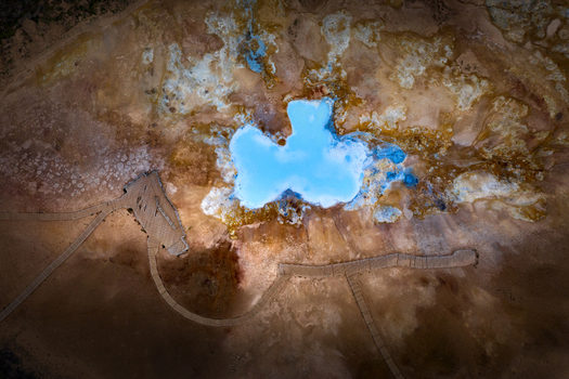 Aerial view of a bright blue geothermal pool surrounded by orange volcanic soil and wooden walkways in Iceland.