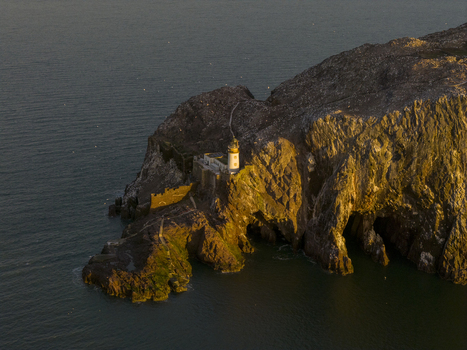 Aerial view of Bass Rock Lighthouse on the steep volcanic cliffs of Bass Rock island with golden sunlight illuminating the stone tower and rocks North Berwick Scotland United Kingdom. Aerial view of Bass Rock Lighthouse on the steep volcanic cliffs of Bass Rock island with golden sunlight illuminating the stone tower and rocks North Berwick Scotland United Kingdom.