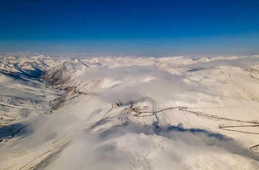 Aerial view of Babusar Pass winding through snow covered mountain peaks under a clear blue sky with low clouds Babusar KPK Pakistan.