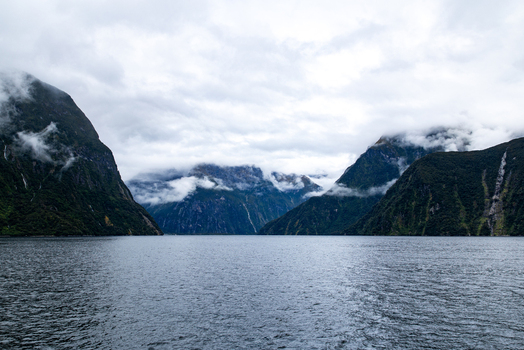 View of majestic dark mountains rise from the serene rippled waters under a blanket of misty clouds Milford Sound Southland Region New Zealand.