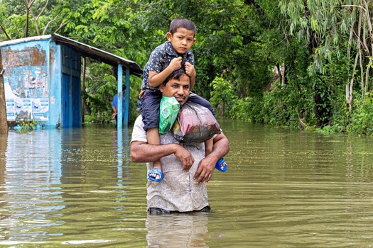Banshkhali Upazila Chittagong Division Bangladesh 10 July 2026 View of a man carrying a child on his shoulders through floodwaters with lush trees and a blue structure in the background. Banshkhali Upazila Chittagong Division Bangladesh 10 July 2026 View of a man carrying a child on his shoulders through floodwaters with lush trees and a blue structure in the background.