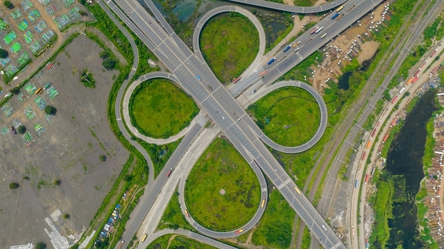 Aerial view of the JNPT Interchange featuring complex cloverleaf ramps surrounding green spaces and nearby industrial storage areas in Navi Mumbai Maharashtra India.