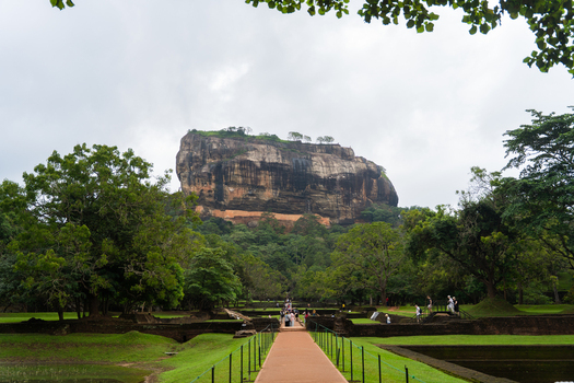 View of the ancient Sigiriya Rock fortress rising above lush gardens and stone ruins with tourists walking along the path in Sigiriya Central Province Sri Lanka. View of the ancient Sigiriya Rock fortress rising above lush gardens and stone ruins with tourists walking along the path in Sigiriya Central Province Sri Lanka.