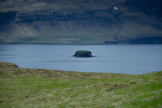 View of Hvalfjordur fjord featuring a small rocky island in the calm water with steep mountains and green grassy hills Iceland. View of Hvalfjordur fjord featuring a small rocky island in the calm water with steep mountains and green grassy hills Iceland.