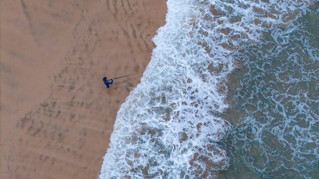 Aerial view of a lone fisherman standing on the sandy shore of Hartenbos beach with white waves crashing against the coast Mossel Bay Western Cape South Africa. Aerial view of a lone fisherman standing on the sandy shore of Hartenbos beach with white waves crashing against the coast Mossel Bay Western Cape South Africa.