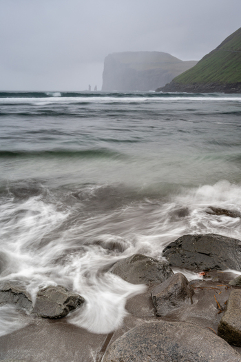 View of the Atlantic Ocean waves crashing against dark volcanic rocks on a misty coastline with distant sea stacks and green cliffs in the Faroe Islands.