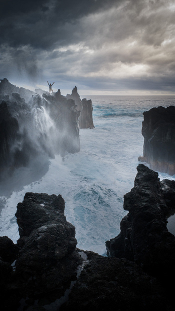 Aerial view of a person standing on volcanic cliffs with arms raised as waves crash against the dark rocks under a dramatic sky in La Reunion Nouvelle Aquitaine France. Aerial view of a person standing on volcanic cliffs with arms raised as waves crash against the dark rocks under a dramatic sky in La Reunion Nouvelle Aquitaine France.