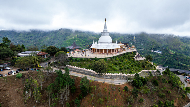 Aerial view of the white Sri Sath Budu Maha Seya stupa perched on a lush green hilltop overlooking the misty mountains of Ella Uva Province Sri Lanka.