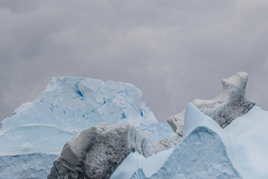View of a large jagged iceberg with deep blue hues and textured ice formations under a cloudy sky in South Orkney Islands. View of a large jagged iceberg with deep blue hues and textured ice formations under a cloudy sky in South Orkney Islands.