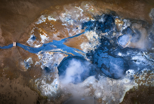 Aerial view of a geothermal field featuring vibrant blue mud pools rising steam vents and mineral rich orange earth across the volcanic landscape of Iceland.