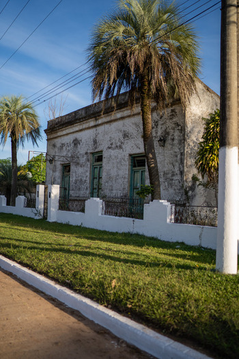View of a weathered historic building with green doors and windows framed by palm trees and a white fence in Guaviravi Corrientes Province Argentina.