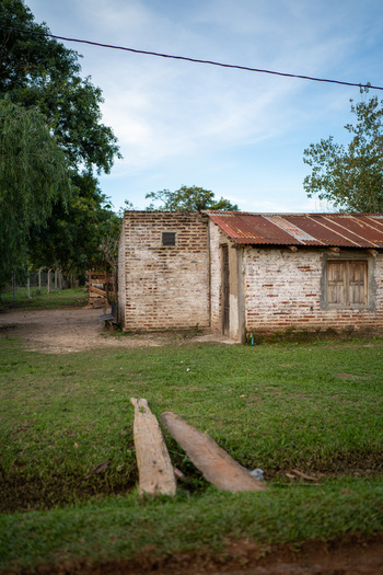 View of a rustic brick house with a weathered corrugated metal roof set in a grassy rural landscape with wooden planks crossing a small ditch in Guaviravi Corrientes Province Argentina.