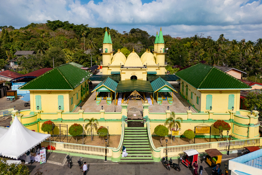 Aerial view of Sultan Riau Grand Mosque featuring its distinctive yellow walls and green tiled roofs under a bright blue sky in Tanjung Pinang Kota Riau Islands Indonesia. Aerial view of Sultan Riau Grand Mosque featuring its distinctive yellow walls and green tiled roofs under a bright blue sky in Tanjung Pinang Kota Riau Islands Indonesia.