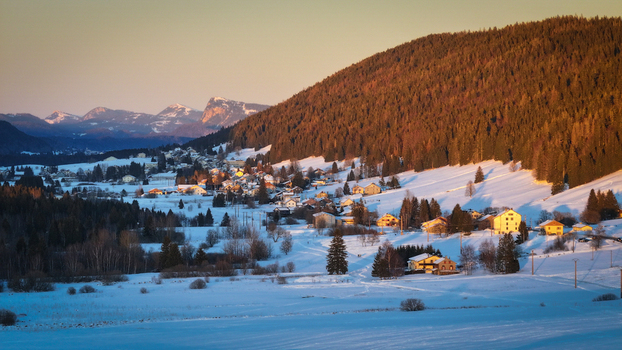 Aerial view of a snow covered village nestled at the foot of a forested mountain bathed in the warm glow of the setting sun Les Rousses Bourgogne Franche Comté France. Aerial view of a snow covered village nestled at the foot of a forested mountain bathed in the warm glow of the setting sun Les Rousses Bourgogne Franche Comté France.
