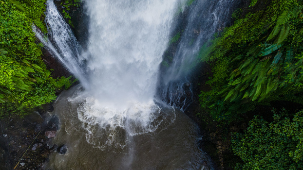 Aerial view of Gomblang Waterfall cascading into a pool surrounded by lush tropical greenery and ferns in Kecamatan Kedungbanteng Jawa Tengah Indonesia. Aerial view of Gomblang Waterfall cascading into a pool surrounded by lush tropical greenery and ferns in Kecamatan Kedungbanteng Jawa Tengah Indonesia.