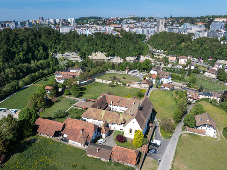 Aerial view of Monastere de Montorge featuring historic stone buildings with red tiled roofs lush green meadows and the urban skyline under a clear blue sky in Fribourg Fribourg Switzerland.