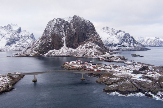 Aerial view of the Lofoten archipelago featuring a bridge connecting islands with red houses surrounded by snow capped mountains and calm sea waters in Nordland Norway.