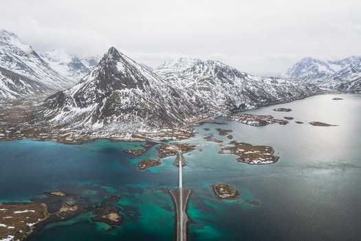 Aerial view of bridges connecting small islands amidst the turquoise waters embraced by snow capped mountains under a cloudy sky Flakstadbruene Nordland Norway. Aerial view of bridges connecting small islands amidst the turquoise waters embraced by snow capped mountains under a cloudy sky Flakstadbruene Nordland Norway.