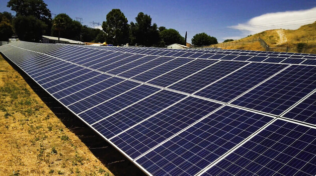 View of a ground mounted solar photovoltaic array in a field with dry grass and trees under a clear blue sky in Los Angeles CA United States.