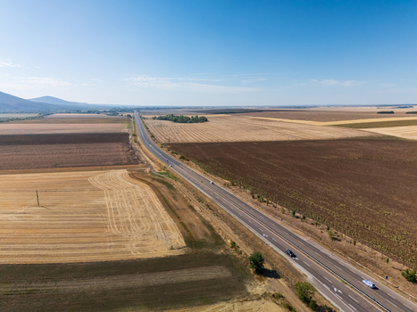 Aerial view of Road 21 near Paszto in Nograd County Hungary with traffic moving through rural farmland fields and meadows beneath a clear summer sky. Aerial view of Road 21 near Paszto in Nograd County Hungary with traffic moving through rural farmland fields and meadows beneath a clear summer sky.