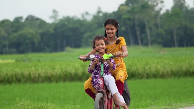 View of two young girls riding a bicycle through rural fields Bangladesh.