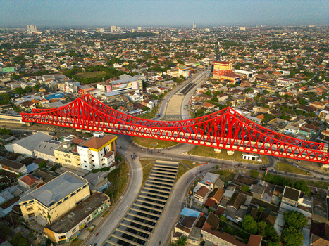 Aerial view of the red steel truss of Jembatan Kereta Api Joglo spanning over an urban road intersection and underpass surrounded by city buildings Kecamatan Banjarsari Jawa Tengah Indonesia.
