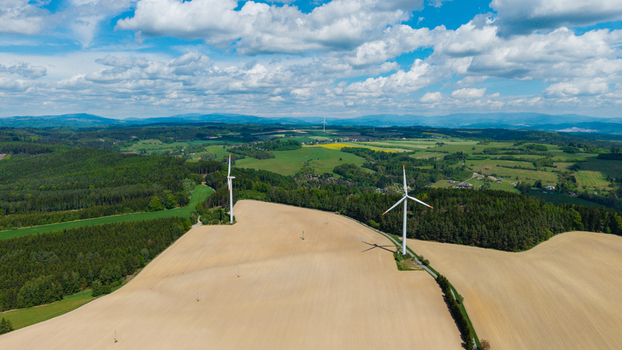 Aerial view of Zipotin wind turbines standing in a vast agricultural field surrounded by dense green forests and rolling hills under a blue sky in Gruna Pardubicky kraj Czechia. Aerial view of Zipotin wind turbines standing in a vast agricultural field surrounded by dense green forests and rolling hills under a blue sky in Gruna Pardubicky kraj Czechia.
