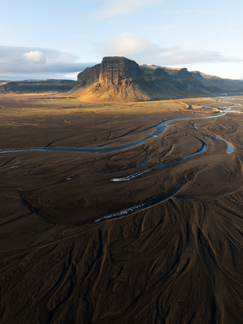 Aerial view of Lomagnupur mountain rising above the vast dark volcanic sand plains and winding glacial river channels under soft golden hour light in Skaftarhreppur Iceland.