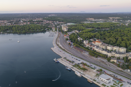 Aerial view of the coastal harbor area in Pula featuring docked boats railway tracks along the waterfront and urban buildings with trees. Pula Istarska zupanija Croatia. Aerial view of the coastal harbor area in Pula featuring docked boats railway tracks along the waterfront and urban buildings with trees. Pula Istarska zupanija Croatia.